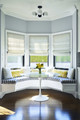 A curved upholstered seat cushion in Capri Outdoor Woven Azzurro fabric anchors this dining area, complementing the geometric abstraction of Cremaillere Soft Blue blinds that frame the bay window. The playful interplay of shapes and soft hues creates a serene yet dynamic space, inviting you to relax and take in the moment.